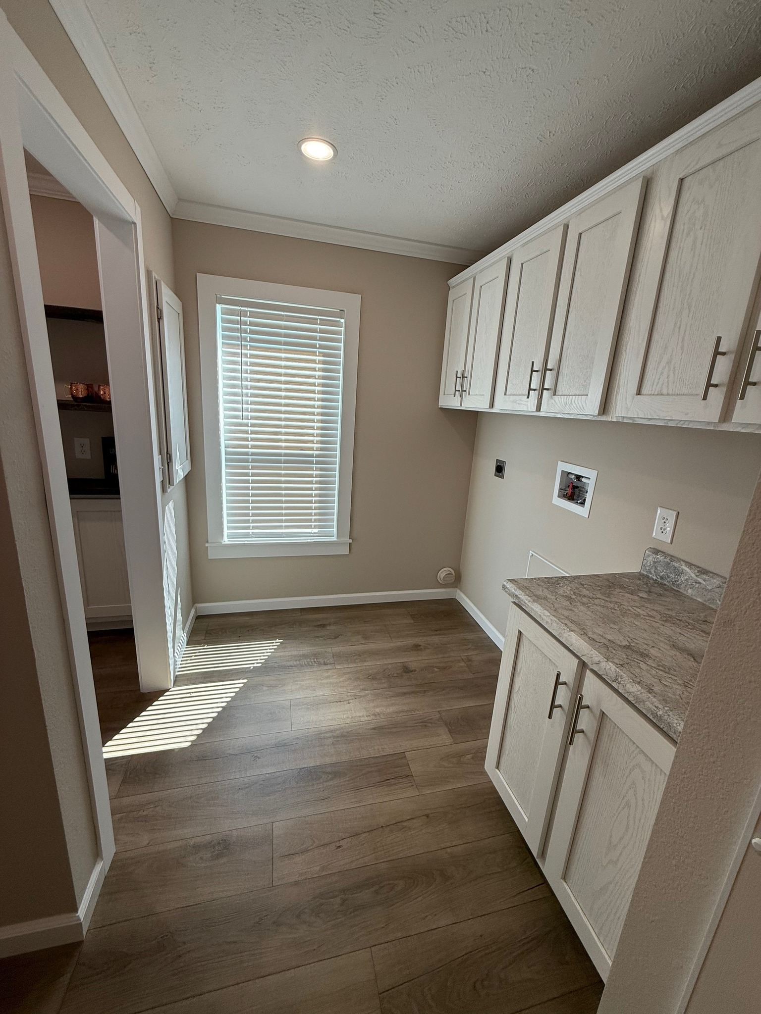Bright laundry room with light beige walls, wood flooring, and a window with blinds. White cabinets and a marble countertop offer storage space.