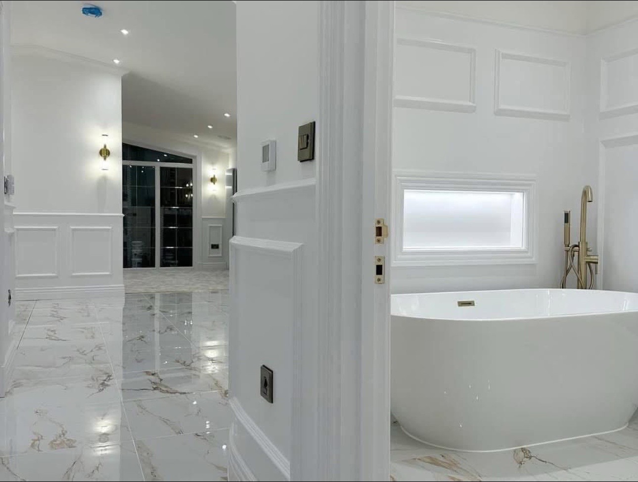 Luxurious bathroom interior with marble floors, white walls, and elegant lighting. Features a large white bathtub and gold fixtures for a modern touch.