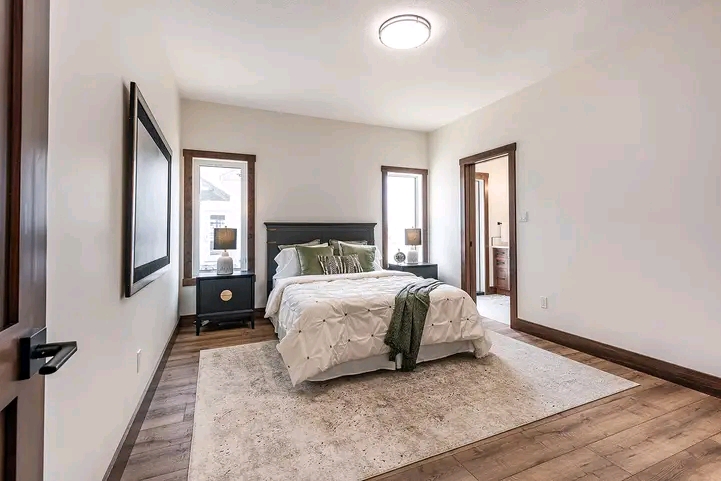 Cozy bedroom with wooden floors, a plush bed with green and white bedding, two nightstands with lamps, bright windows, and an adjacent bathroom door.