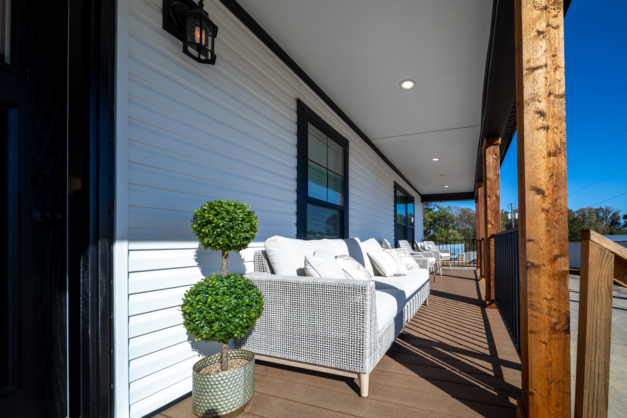 A sunny porch with white siding, featuring a wicker couch with white cushions and a round potted plant. Wooden beams and a black light fixture add contrast.