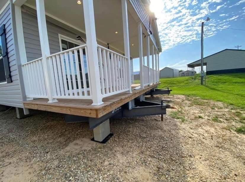 A mobile home sits on large steel beams, with a wooden porch featuring white railings. The setting is a sunny day with scattered clouds and grassy surroundings.