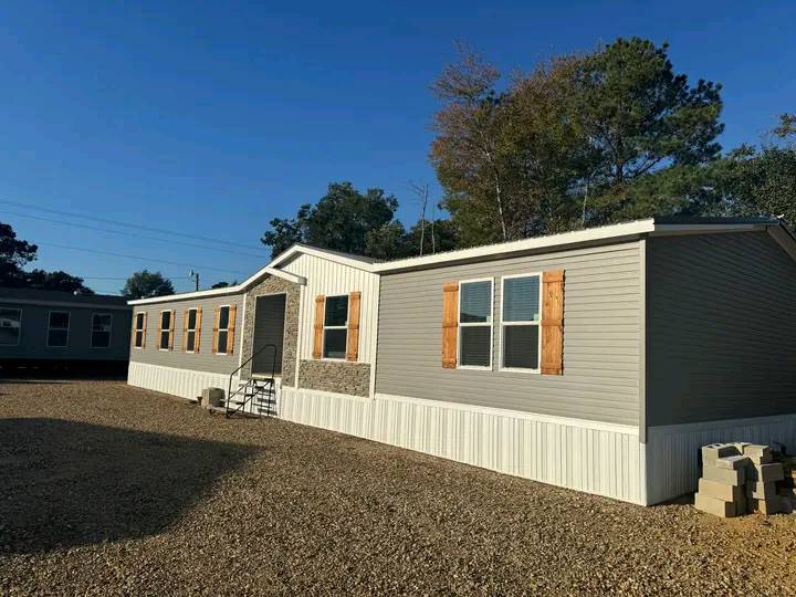 A single-story, light gray mobile home with a white base and wooden shutters sits on a gravel lot. It's sunny, with trees on one side.