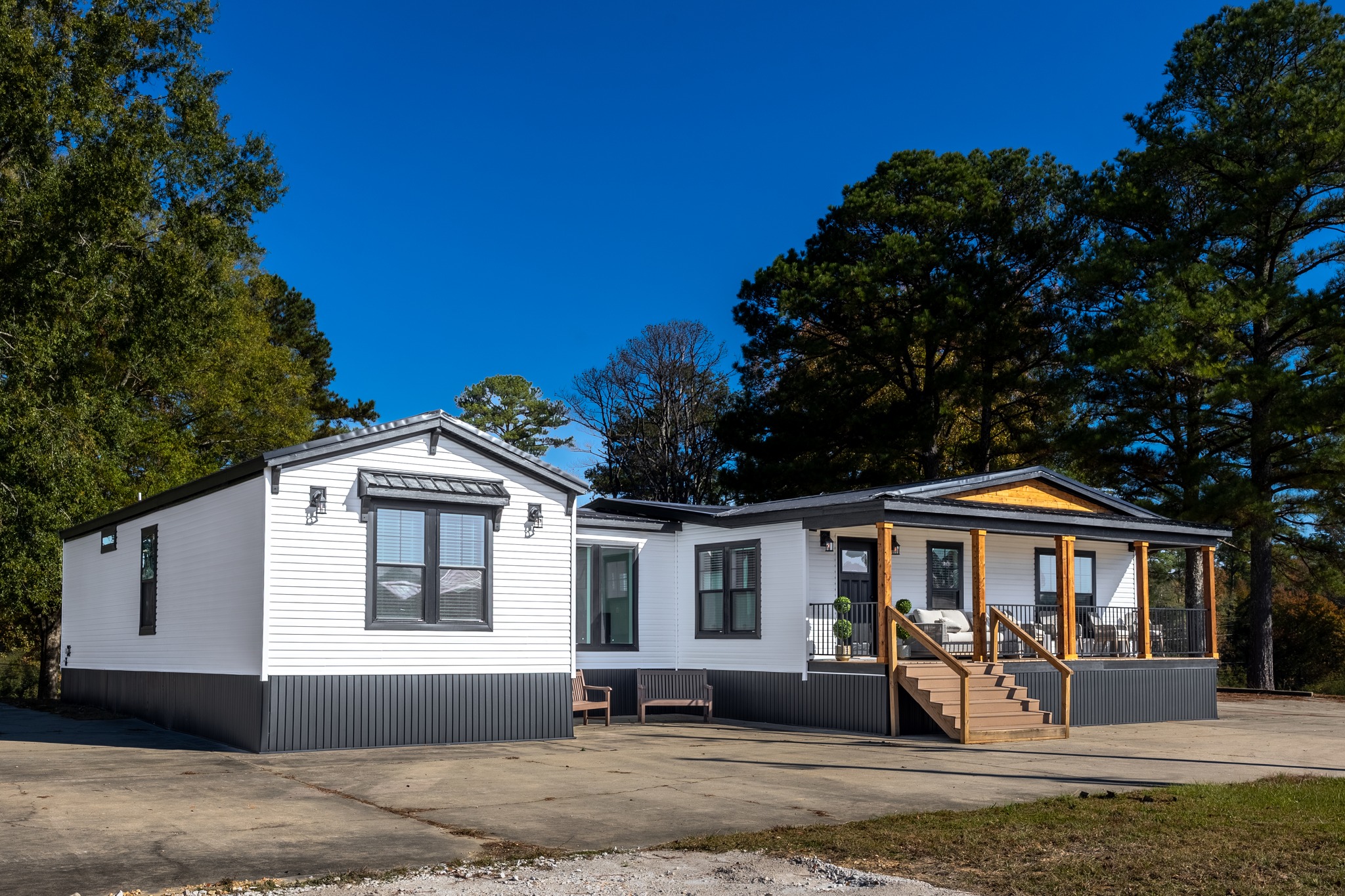 A modern white house with gray trim sits amidst tall trees under a clear blue sky. It features a welcoming porch with wooden steps, exuding a serene vibe.