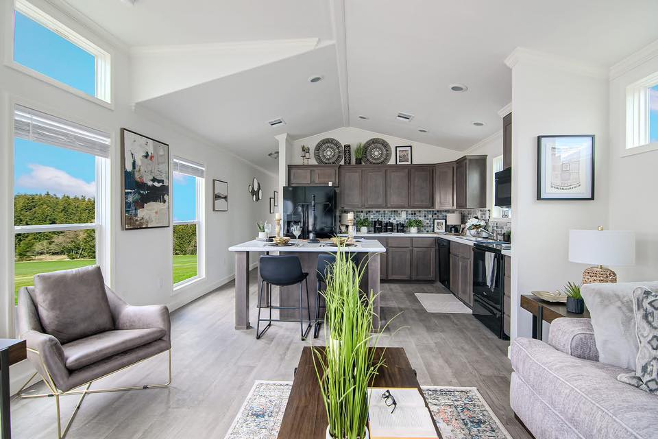 Modern living room and kitchen with high ceilings and large windows overlooking green scenery. Neutral tones, cozy seating, plants, and framed art create a welcoming atmosphere.
