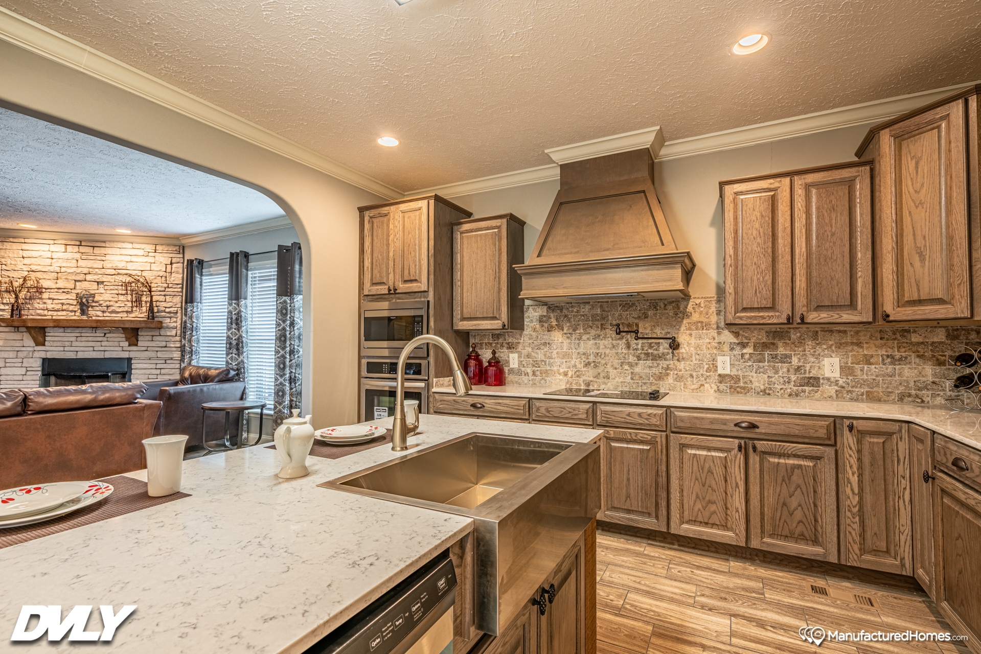 Warm, rustic kitchen with wood cabinets, stone backsplash, and central island featuring a farmhouse sink. Cozy living room with stone fireplace visible through an archway.