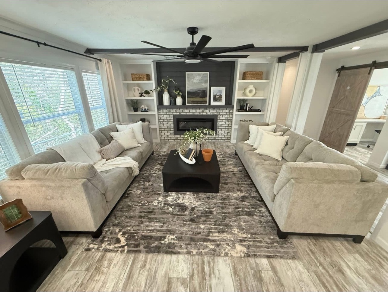 Cozy living room with two beige sofas facing each other, centered around a black coffee table over a textured rug. A brick fireplace with shelves and decor behind, and light streaming in from large windows, creates a warm, inviting atmosphere.