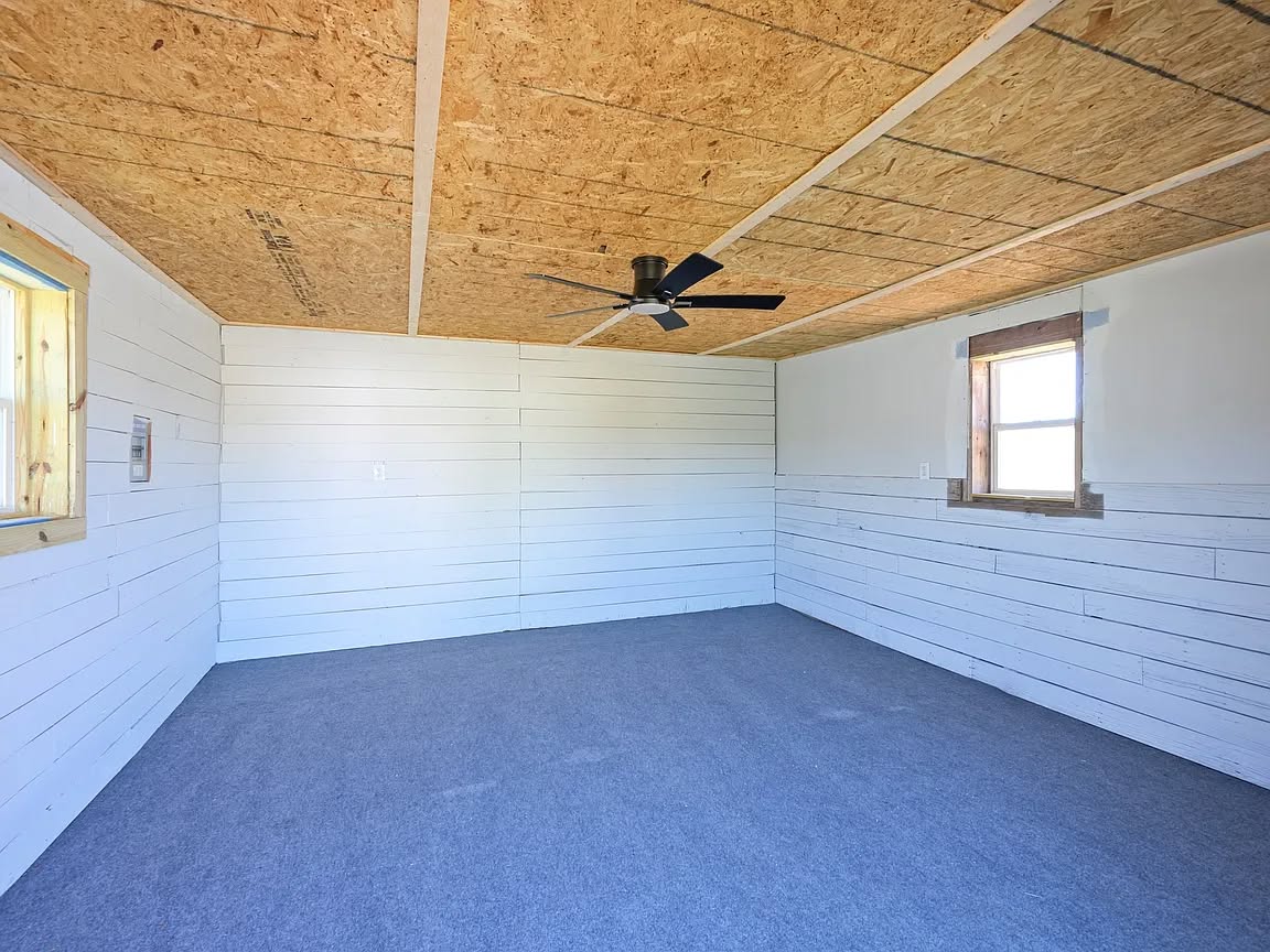 Empty room with white shiplap walls, plywood ceiling, and gray carpet. A black ceiling fan is centered, and a window lets in natural light.