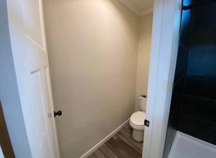 Narrow bathroom with light beige walls and a white toilet against the right wall. Wooden floor and partial view of a black-tiled shower on the right.
