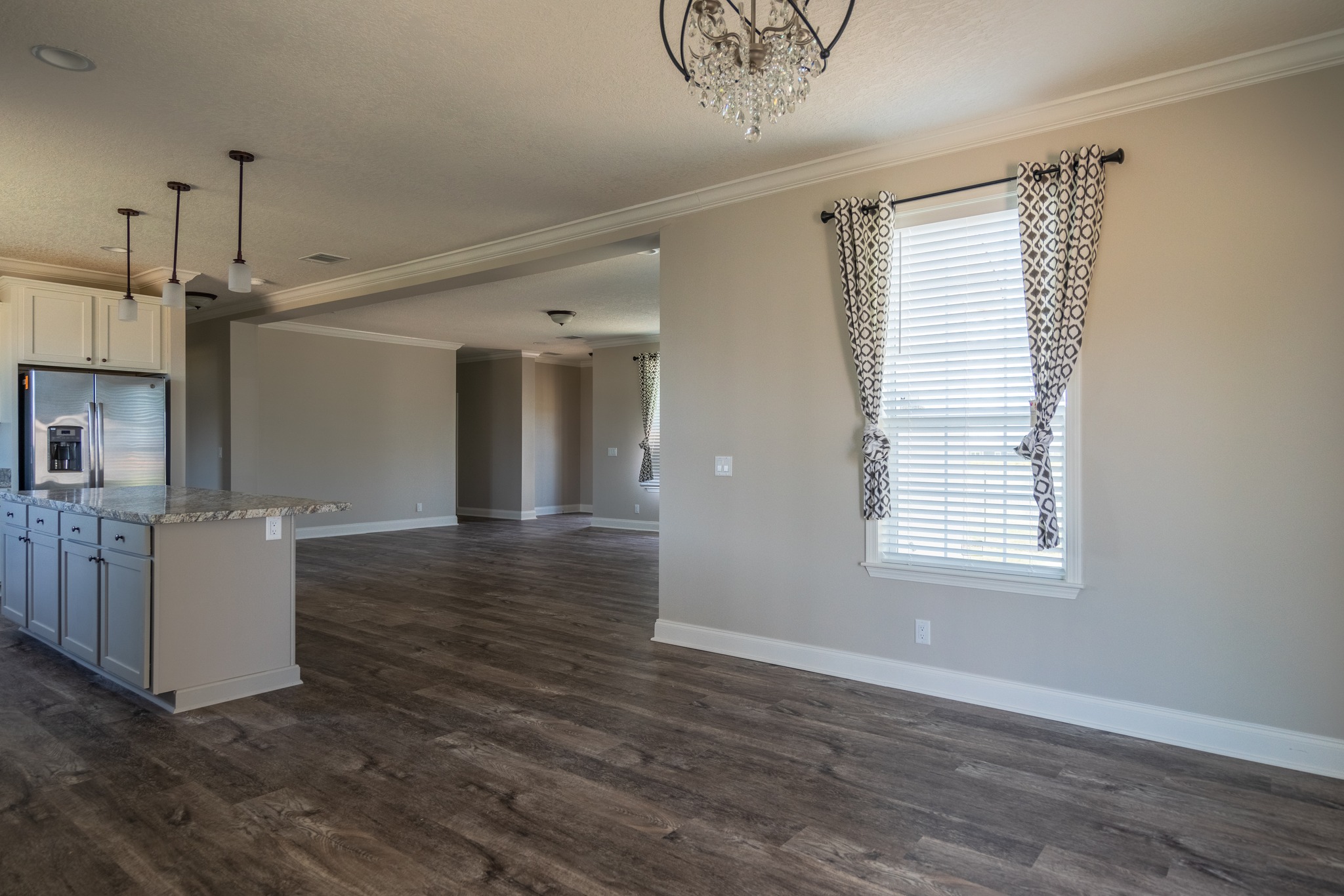 Spacious, empty living area with wood floors and beige walls. Features a modern chandelier, patterned curtains, and a kitchen with granite countertops.
