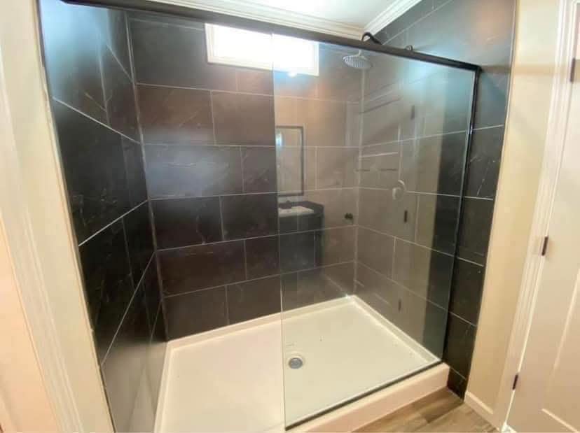 Modern shower with clear glass doors and dark tile walls. Overhead showerhead visible. Bright lighting and neutral tones create a clean, sleek look.