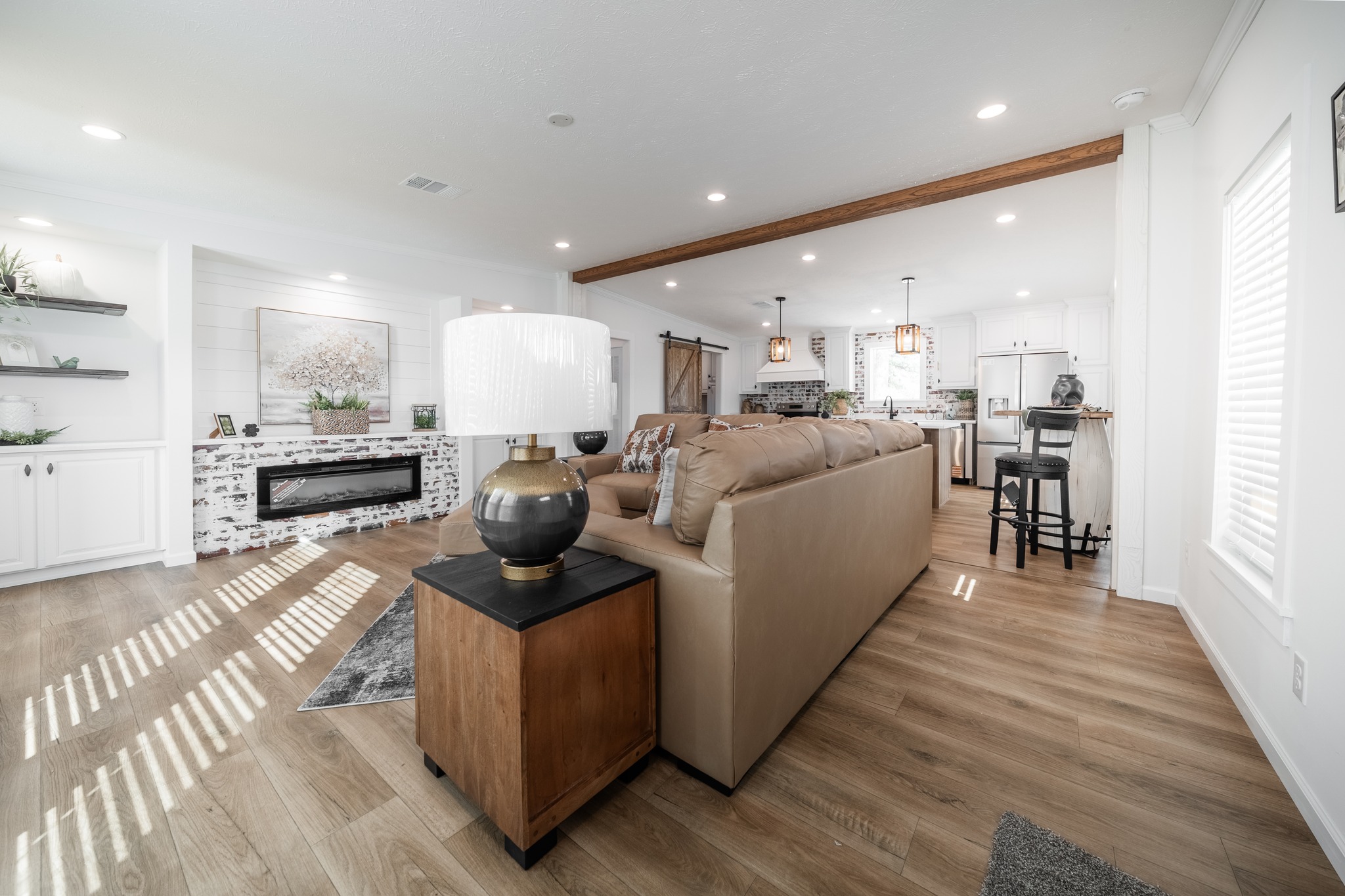 Spacious living area with a tan sofa, wooden floors, and modern decor. Brick-accented fireplace, open kitchen with bar stools, and large table lamp. Cozy ambiance.