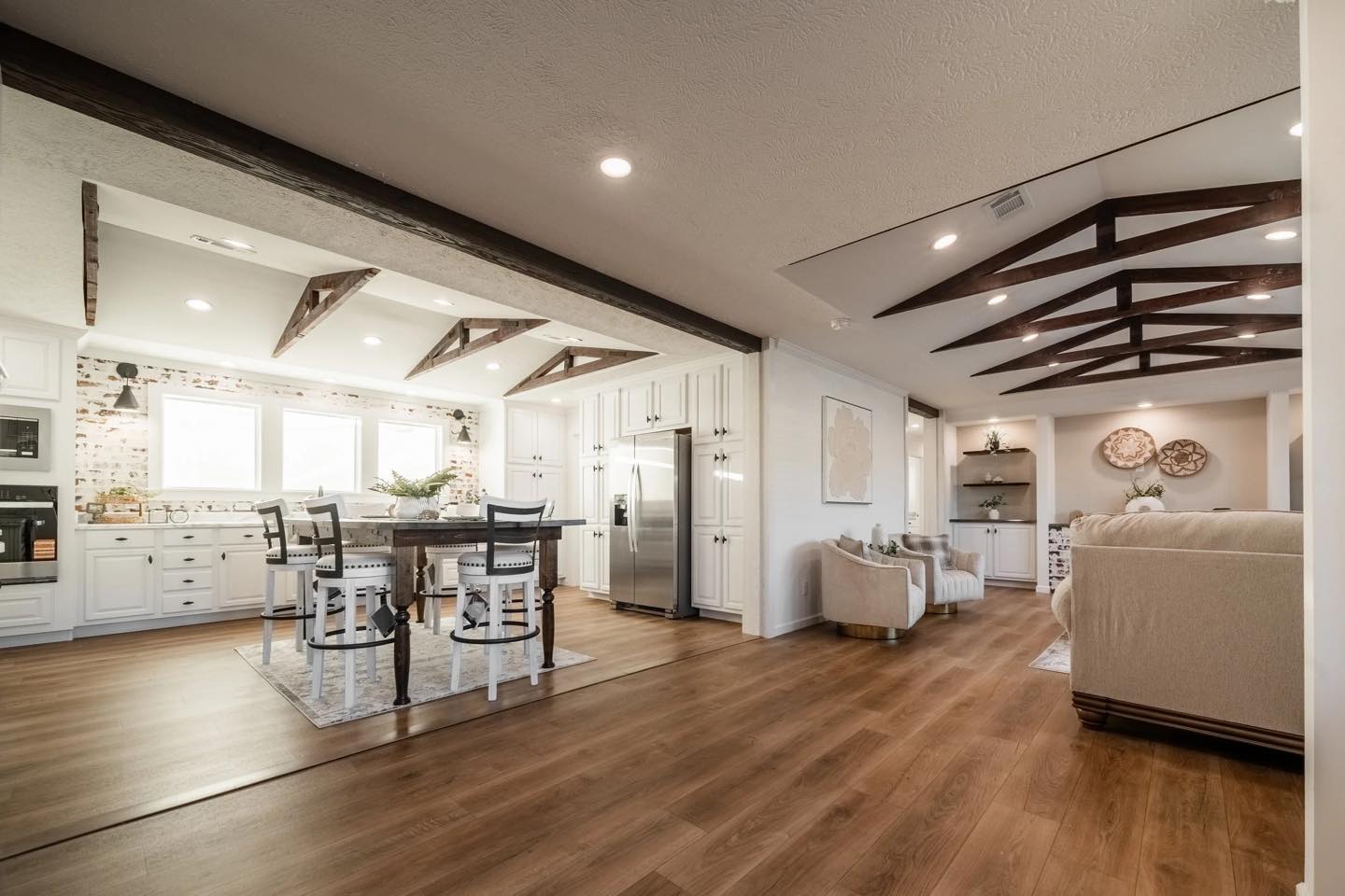 Spacious modern kitchen and living area with wooden floors and exposed beams. White cabinetry, stainless steel appliances, and cozy seating create a warm, inviting atmosphere.