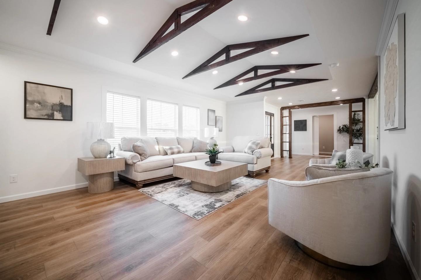 Modern living room with white sofas arranged around a wooden coffee table, under dark wooden ceiling beams. Soft lighting and light wood flooring add warmth.