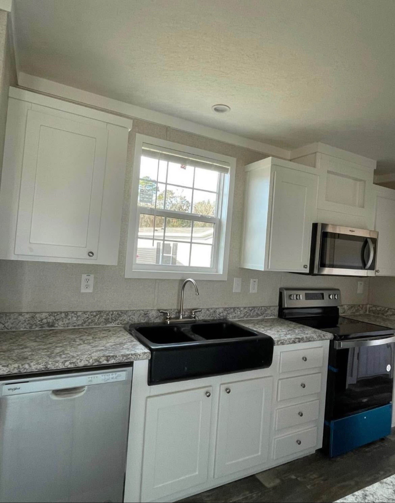 Modern kitchen with white cabinets, marble countertops, and stainless steel appliances. A large window above the black sink provides natural light.