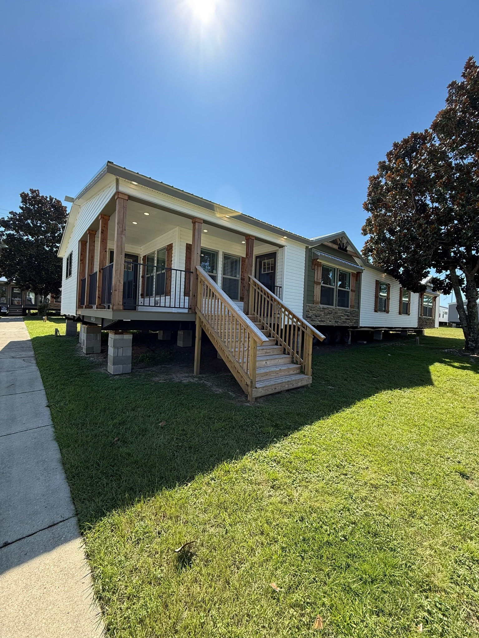 Single-story white house on raised foundation, with wooden porch and steps. Bright sun and clear sky, casting shadows on the green lawn.