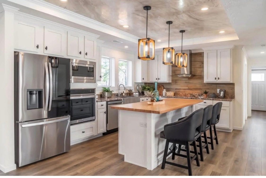 Sleek kitchen with white cabinets, stainless steel appliances, wood accents, and a wood-topped island. Three pendant lights hang above gray chairs.