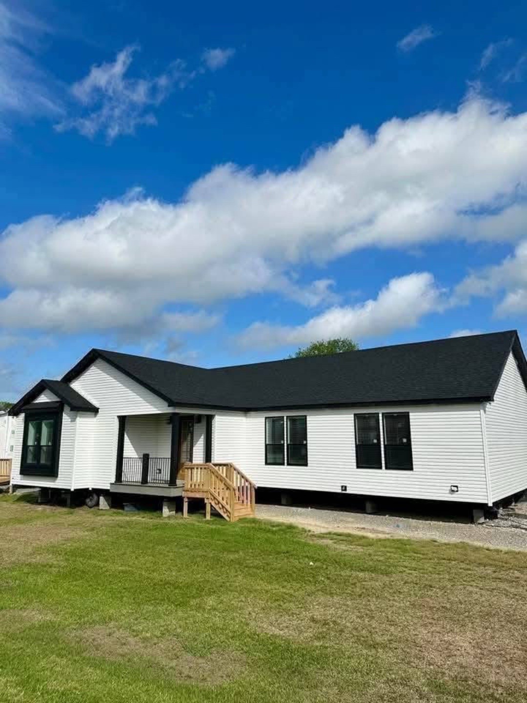 A single-story, white prefabricated house with black roof and windows sits on a grassy lawn under a bright blue sky with scattered clouds.