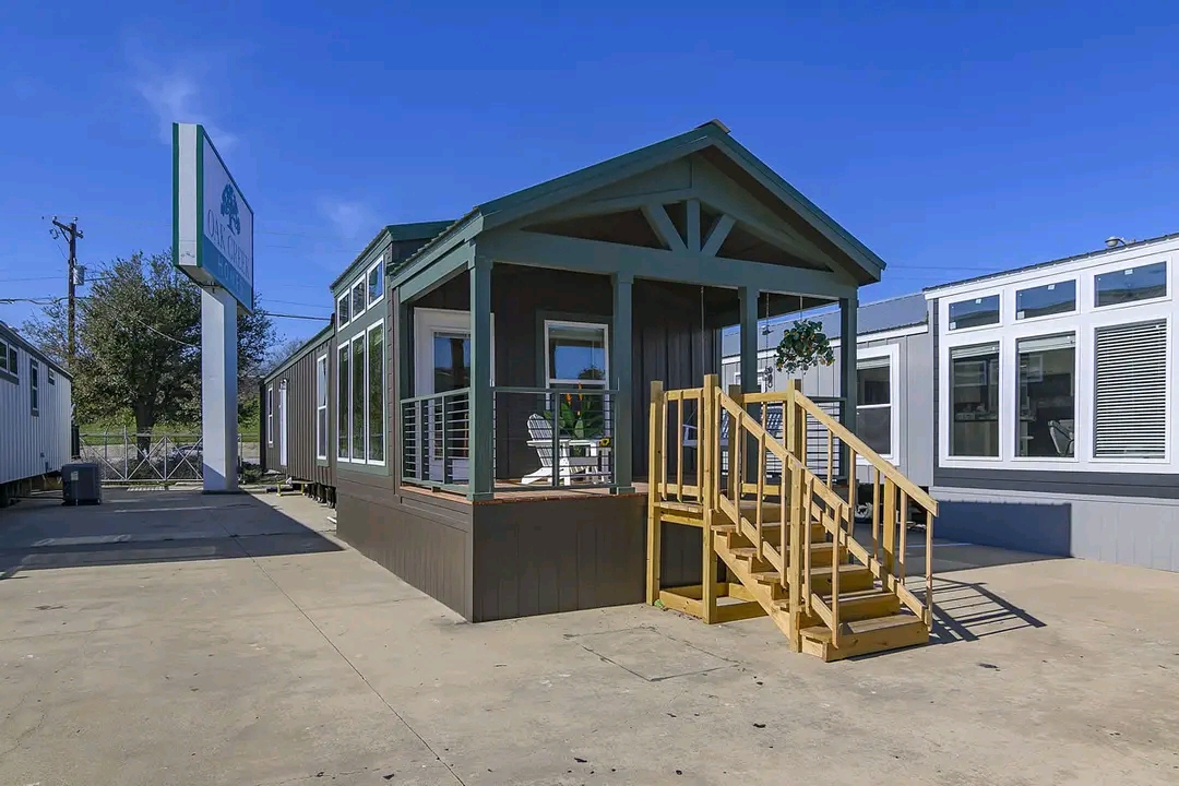 Small modern modular home with a green roof and porch, featuring wooden steps. Clear blue sky and neighboring homes create a calm, inviting scene.