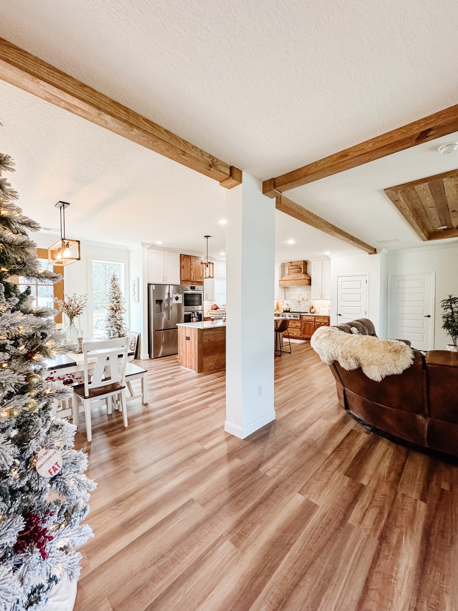 Bright open-concept room with wooden beams features a festive Christmas tree, modern kitchen, and cozy dining area, creating a warm, welcoming atmosphere.