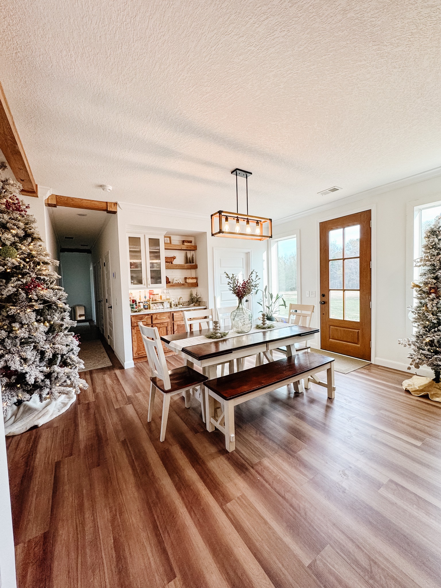 Bright dining area with a wooden table, chairs, and a glass vase centerpiece. Flanked by snowy Christmas trees, a warm, inviting atmosphere prevails.
