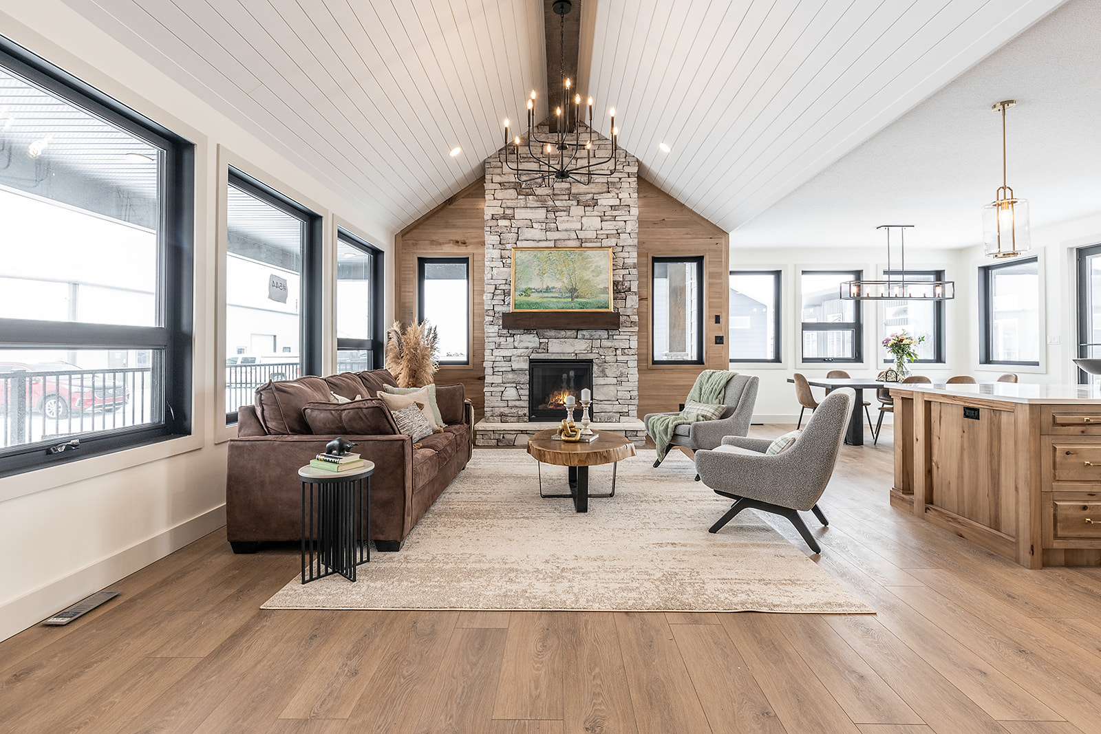 Spacious living room with high white ceilings, stone fireplace, and large windows. Brown leather sofa, cozy chairs, and chic chandelier create a welcoming atmosphere.