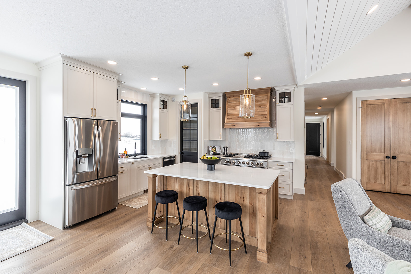Modern kitchen with white cabinets, stainless steel fridge, and wooden accents. An island with three black stools and pendant lights adds elegance.
