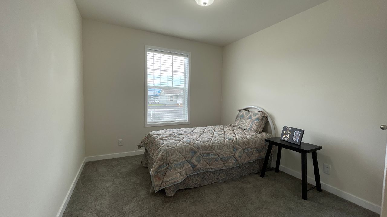 A small, minimalist bedroom features a single bed with a patterned quilt, a window with blinds, and a bedside table holding a small picture frame. The room has a calm, simple atmosphere.