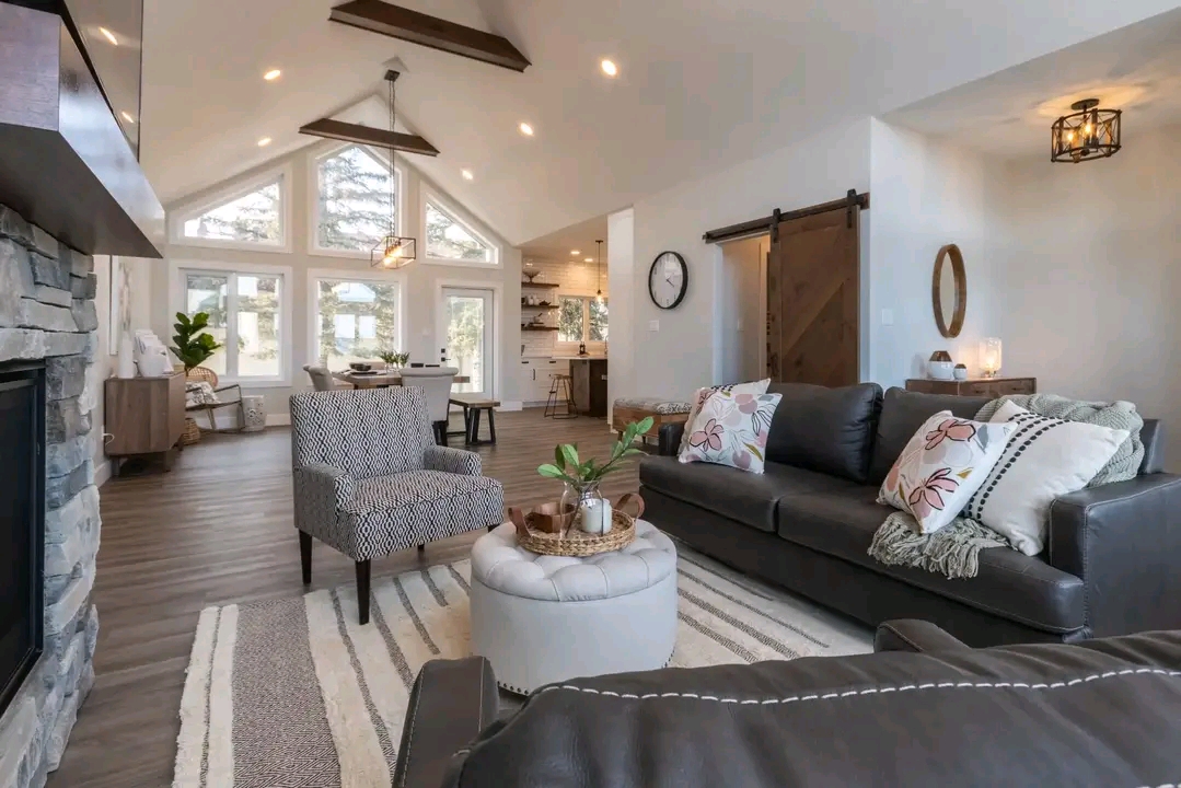 Spacious living room with vaulted ceiling and exposed beams. Features a stone fireplace, gray sofas with floral pillows, rustic decor, and large windows. Cozy and inviting atmosphere.