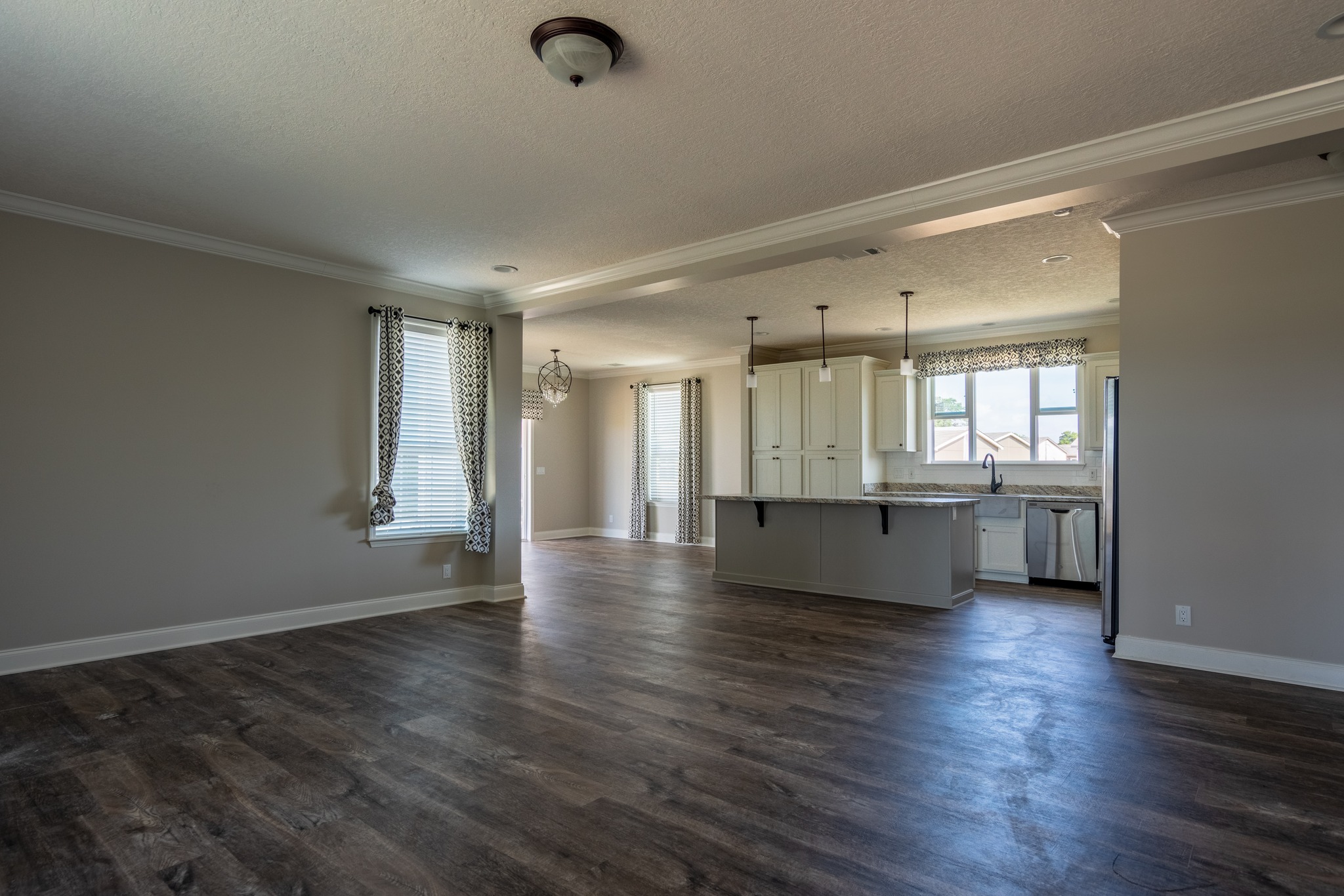 Spacious, empty living room with hardwood floors, beige walls, and a ceiling light. An open kitchen features white cabinets and pendant lighting.
