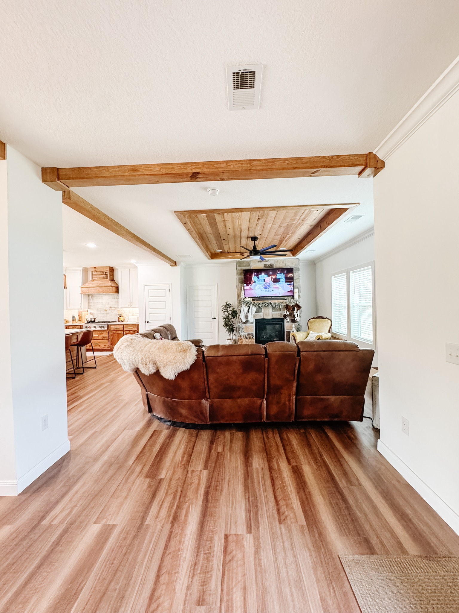 Open-concept living room with wood flooring, brown sectional sofa, and cozy throw. Fireplace with TV above, abundant natural light from large windows.
