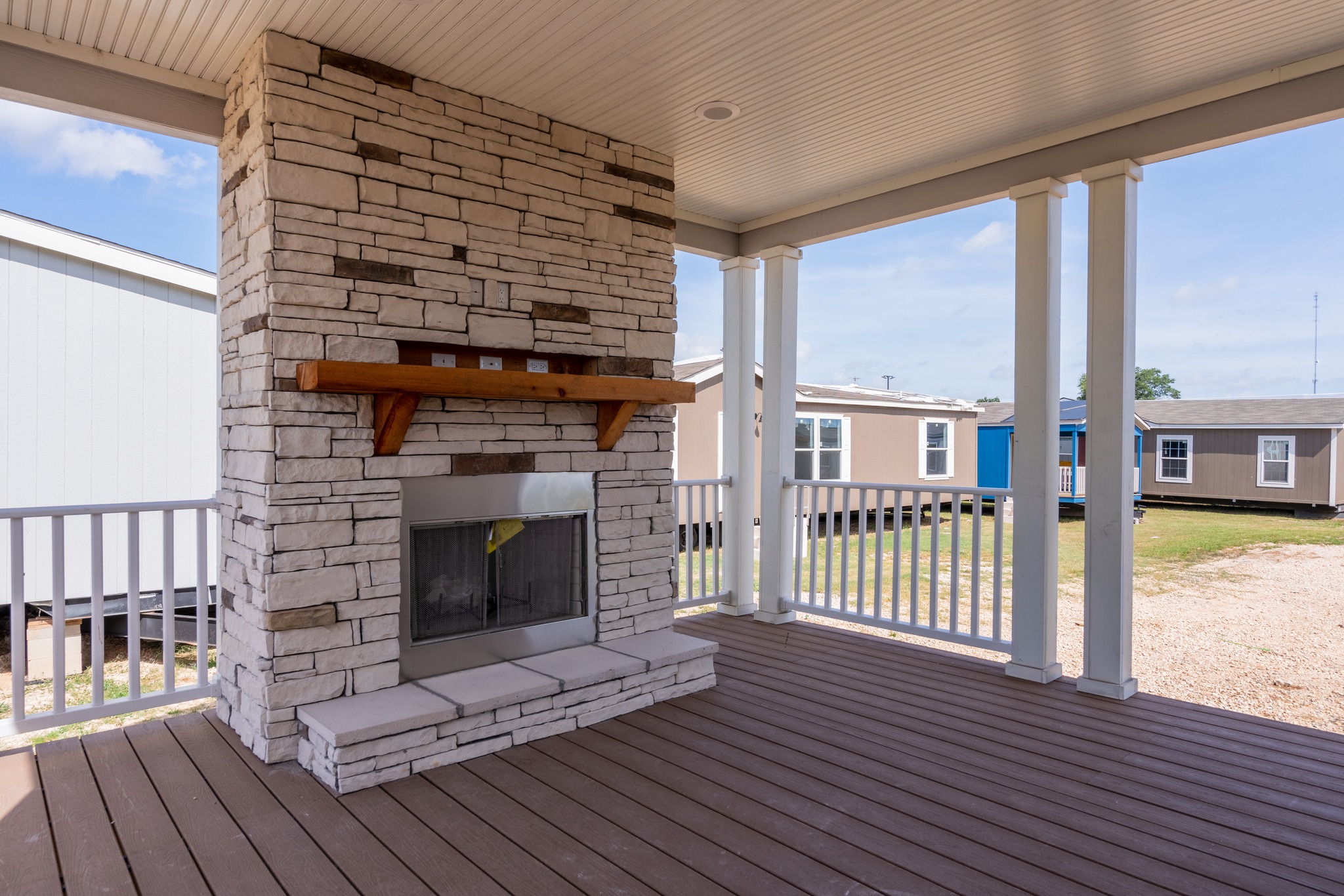 Covered patio with a stone fireplace, wooden mantle, and white railings. Surrounding area shows modular homes under a bright, partly cloudy sky.
