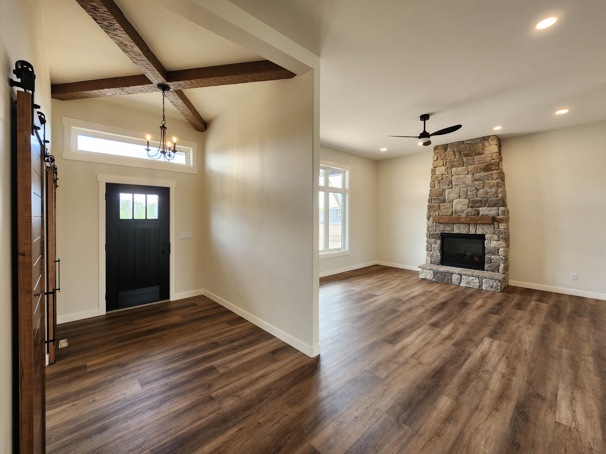 Spacious living area with a stone fireplace and wooden floors. Features a vaulted ceiling with wood beams and a modern chandelier near the entrance.