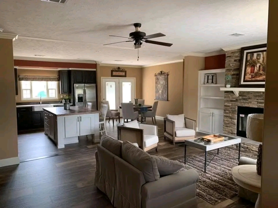 Modern living room with a brown couch, white chairs, stone fireplace, and ceiling fan. Open kitchen with dark cabinets and a dining area in view. Cozy and elegant atmosphere.