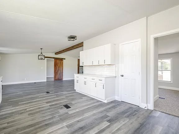 Spacious, modern open-plan room with light gray wood floors, white cabinets, and a wooden beam. Features a sliding barn door and ample natural light.