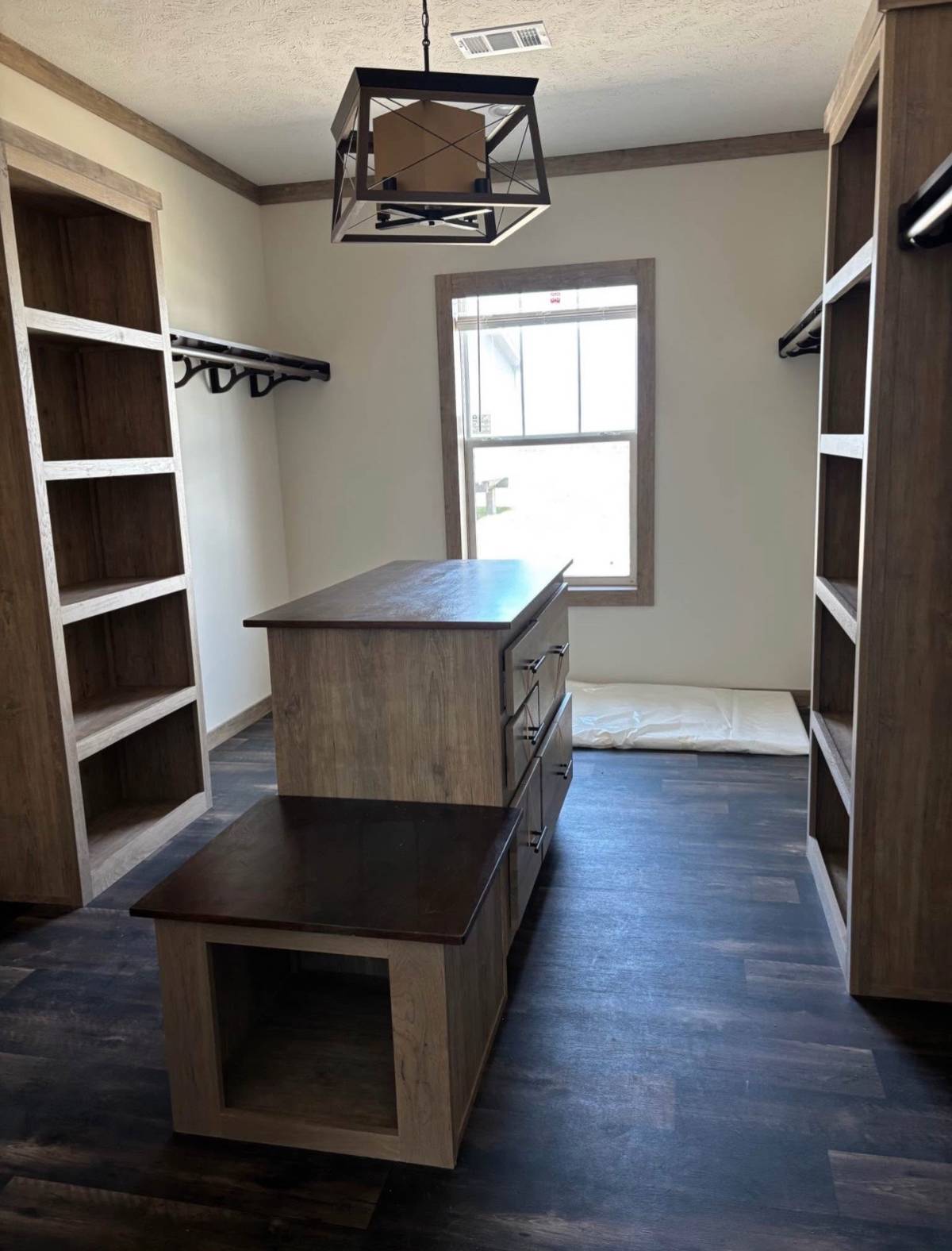 A spacious walk-in closet with dark wood floors and light wood shelves. A central island with drawers stands beneath a modern geometric light fixture.