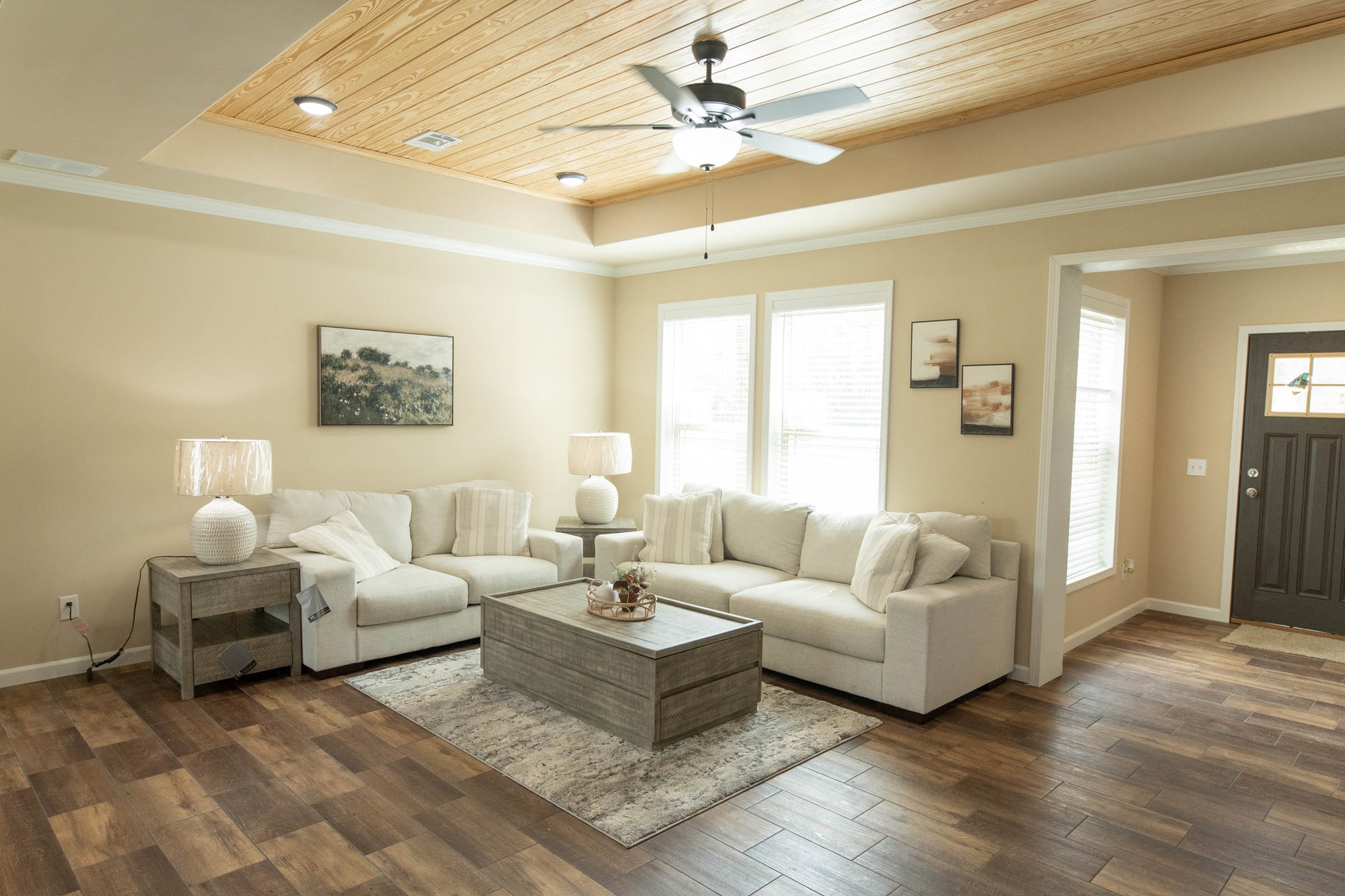 Modern living room with cream sofas, wooden coffee table, and side tables with lamps. Light wood ceiling, ceiling fan, and wide windows create a cozy, elegant ambiance.