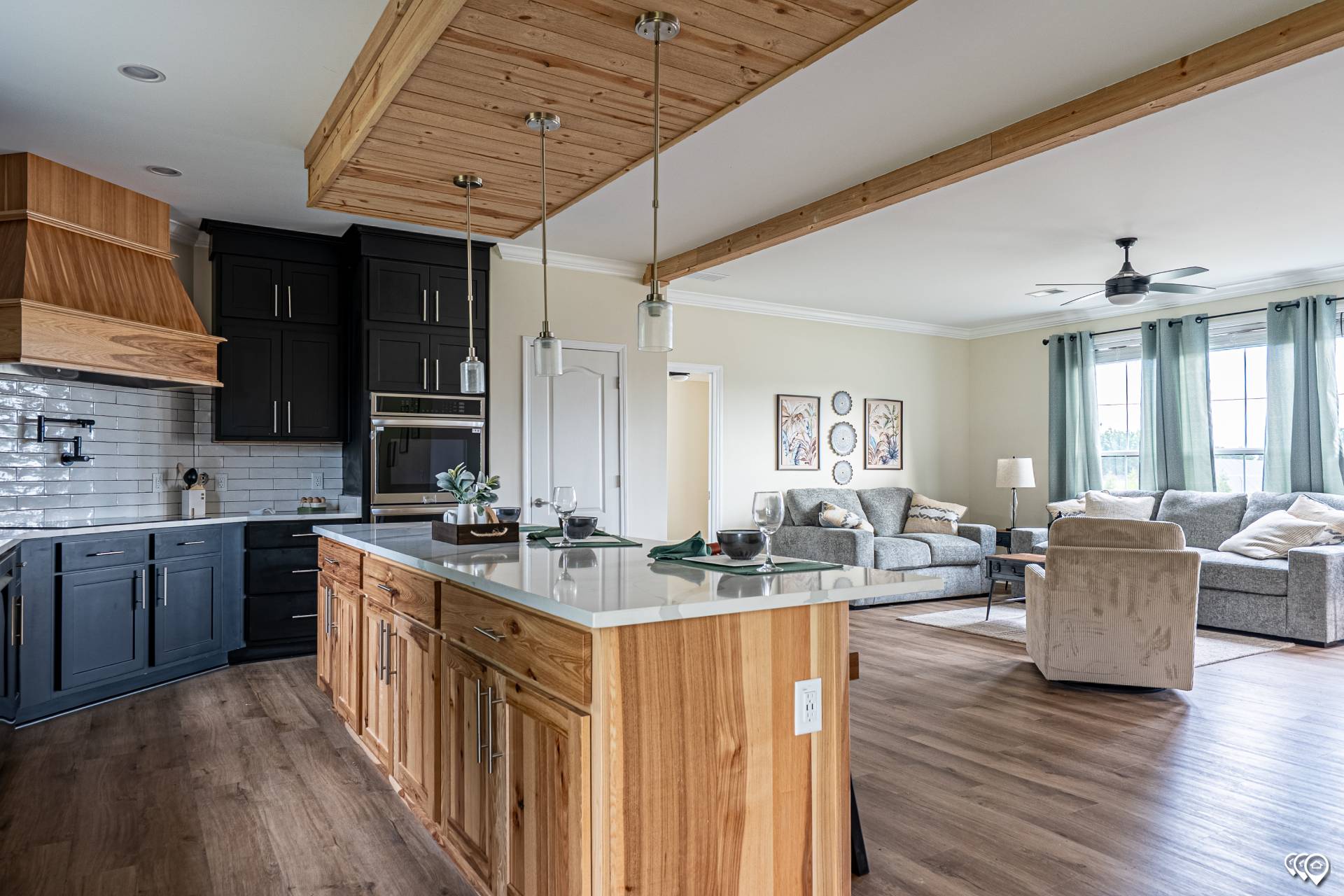 Modern open-concept kitchen and living room with wooden accents. Features dark cabinetry, white countertops, cozy gray seating, and large windows with green curtains.