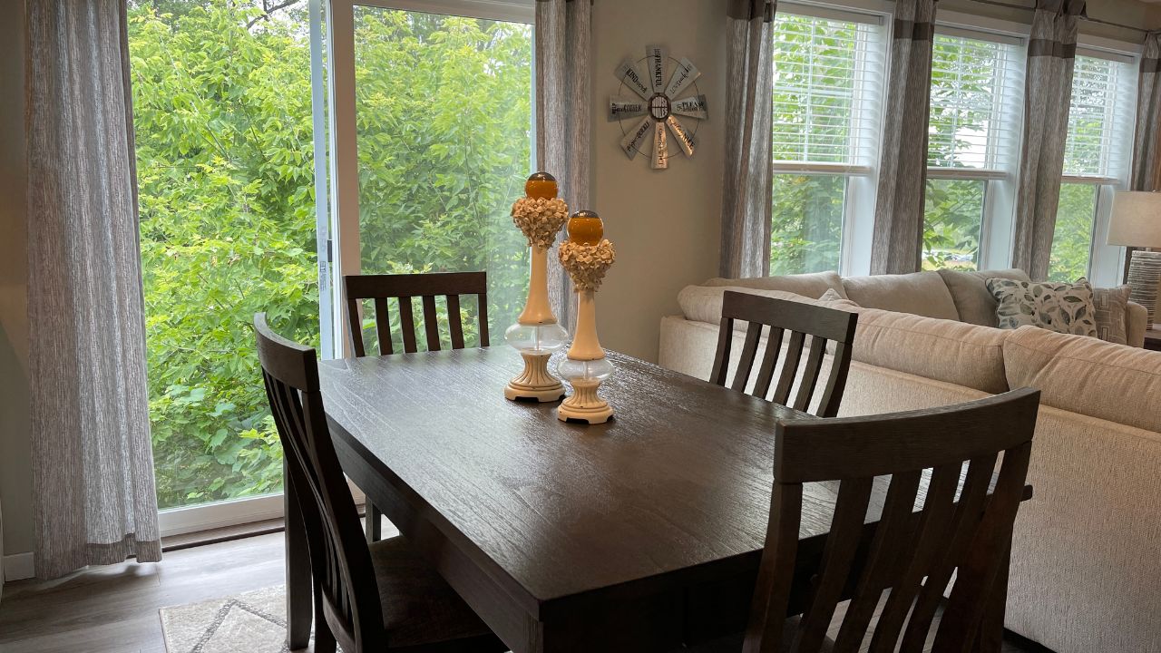 A cozy dining area with a dark wooden table and chairs, two decorative candle holders centered. Large windows show lush greenery outside, creating a serene atmosphere.