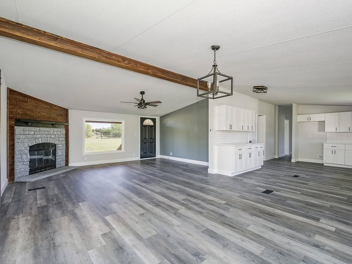 Spacious living room with gray wood flooring, a white stone fireplace, white cabinets, and large window. Neutral tones create an open, airy feel.