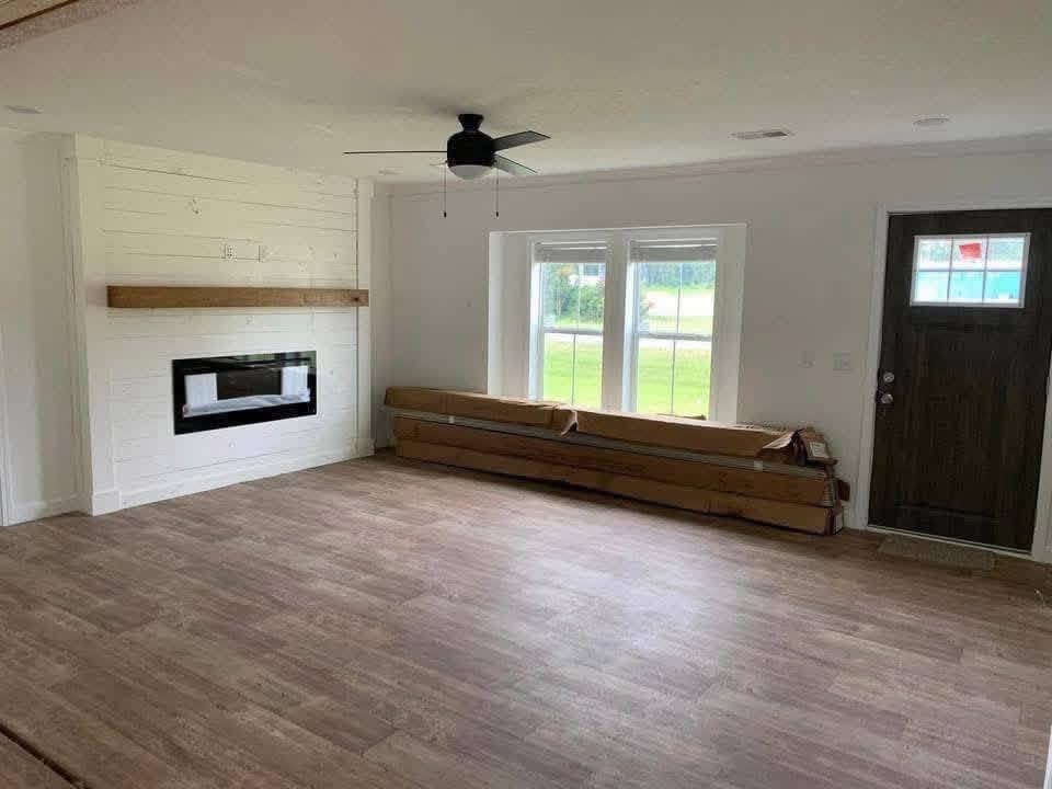 Modern living room with light wood flooring, a sleek fireplace on a white shiplap wall, a large window, rolled carpets, and a dark wood door.