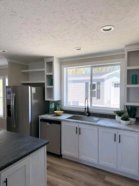 Modern kitchen with white cabinets and gray countertops. Large window over sink with plants and bowls. Natural light creates a fresh, clean ambiance.