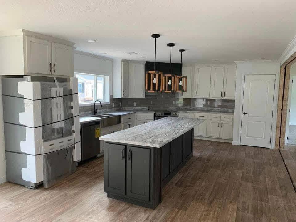 Modern kitchen with a large gray island, wooden floor, gray cabinets, and metallic appliances wrapped in protective film. Three hanging wooden pendant lights.