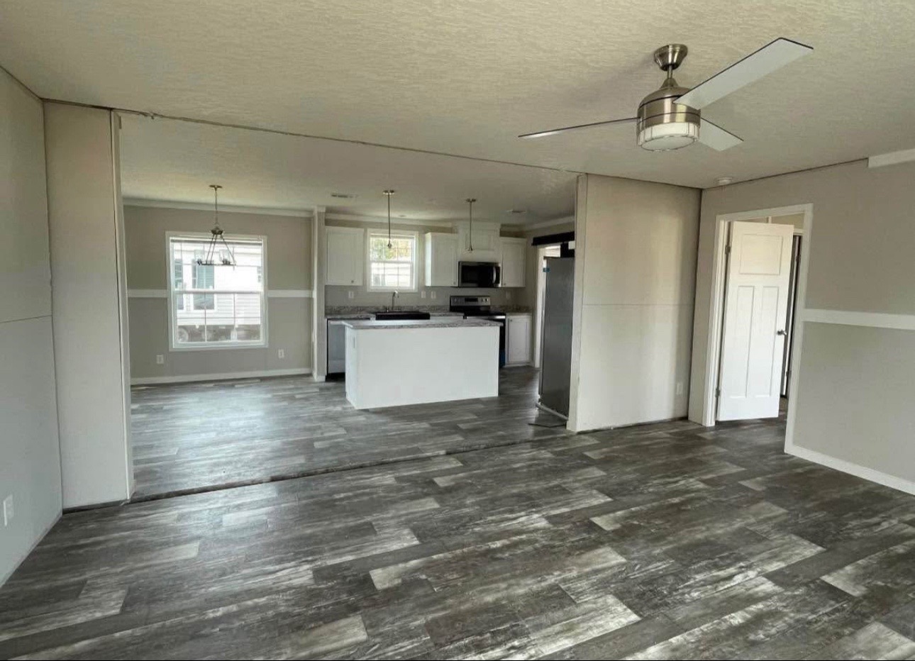 Spacious modern kitchen with gray wood floors, white cabinetry, and a central island. Ceiling fan and large windows create a bright, airy atmosphere.