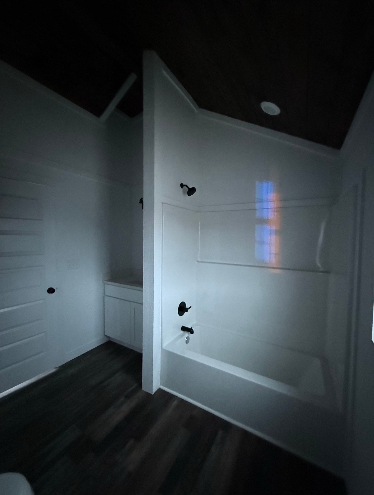 Dimly lit bathroom with dark wooden floors, a white tub, and black fixtures. A faintly illuminated window casts soft light onto the walls.