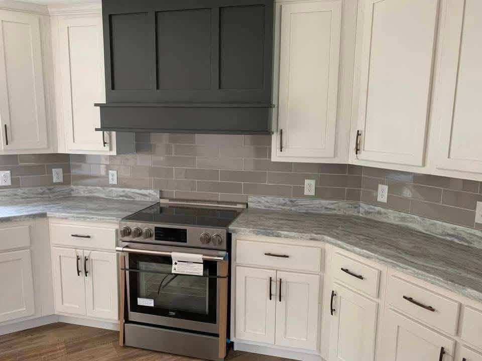 Modern kitchen with light gray cabinets and marble countertops. A stainless steel oven sits below a dark gray range hood. Light gray tile backsplash.