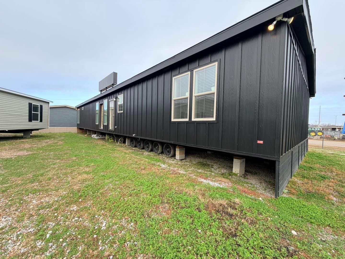 A black modular home on wheels sits on a grassy lot, with overcast skies. The structure features two windows and a simple, modern design.