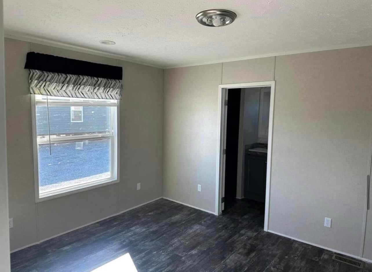 Small room with light gray walls and dark wood flooring. A large window with a zigzag-patterned valance lets in natural light. A door leads to another room.