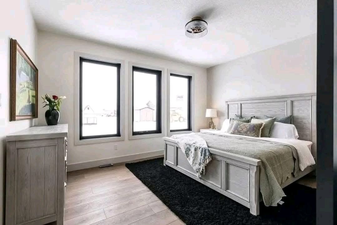 A bright bedroom features a large bed with a plush headboard, a cozy blanket, and patterned pillows. Three tall windows let in natural light, creating an airy feel. A black rug contrasts with light wood flooring. A vase with flowers sits on a dresser, adding a touch of color. The tone is peaceful and inviting.