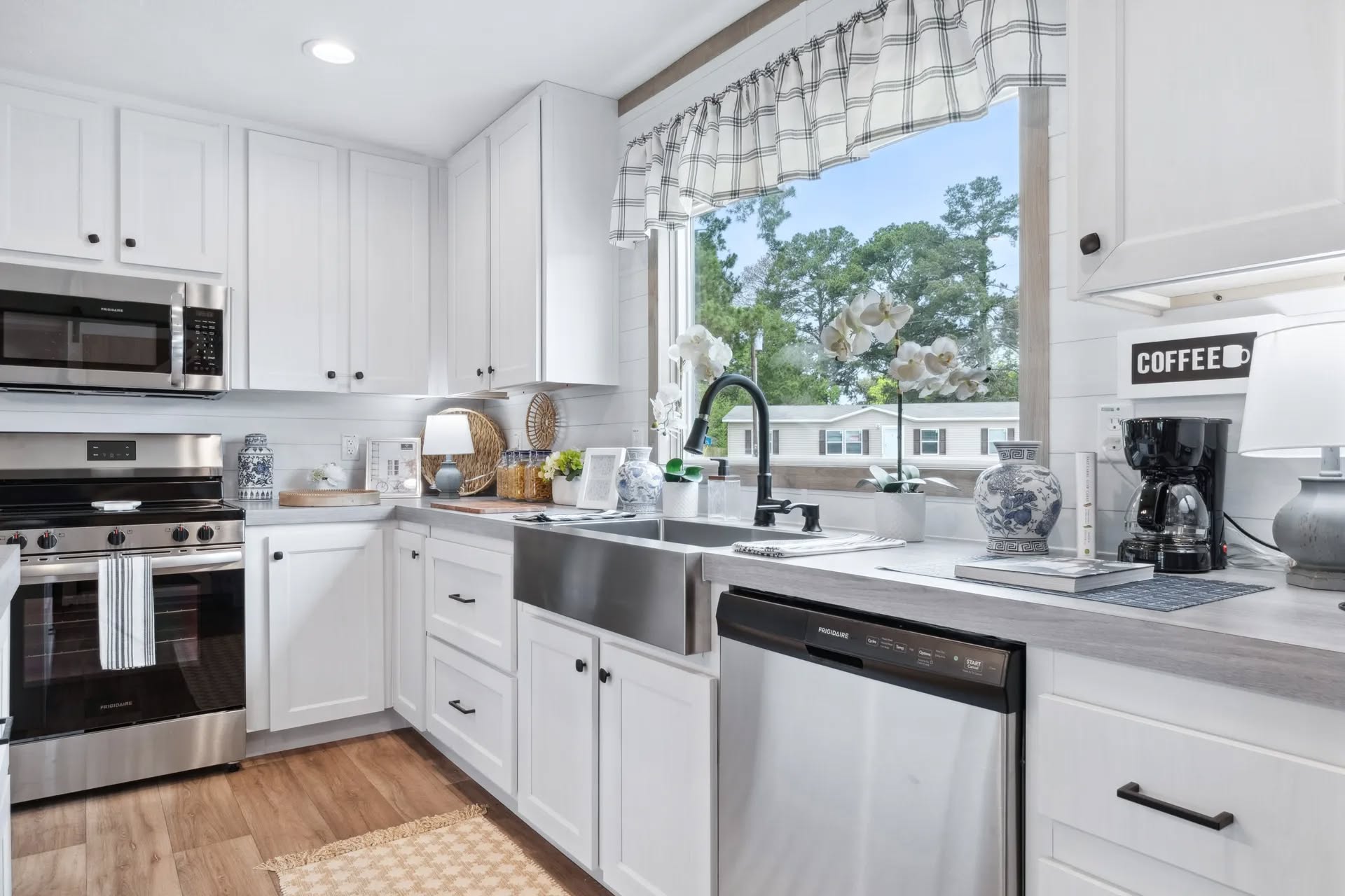 A bright kitchen with white cabinets, stainless steel appliances, and a farmhouse sink beneath a window with checkered curtains. Modern, clean, and inviting.