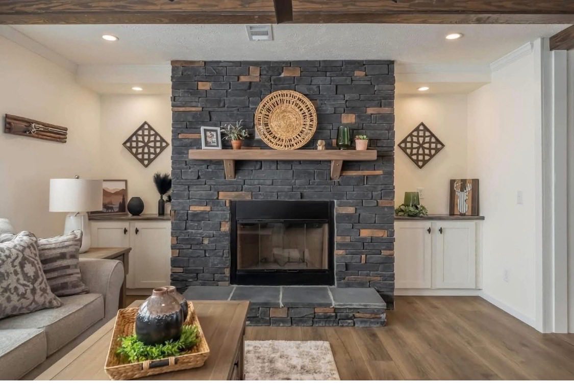Cozy living room with a dark stone fireplace, wooden mantle adorned with plants and decor. Soft lighting, earth tones, and plush furnishings create warmth.